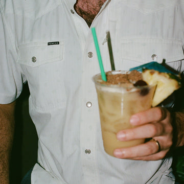 Man wearing The Surf No Turf white button-up shirt holding tropical drink with pineapple garnish