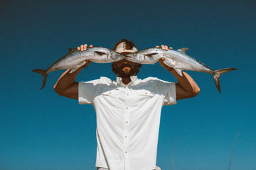 Man in white shirt holding two fish in front of his face against a clear blue sky