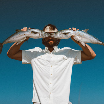 Man wearing The Key West shirt holding two silver fish horizontally in front of face against clear blue sky