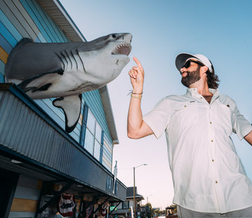 Man in white shirt and cap pointing at large shark sculpture on building exterior at sunset