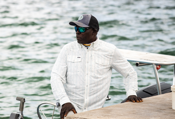Man wearing white long-sleeve fishing shirt and cap leaning on boat by the water
