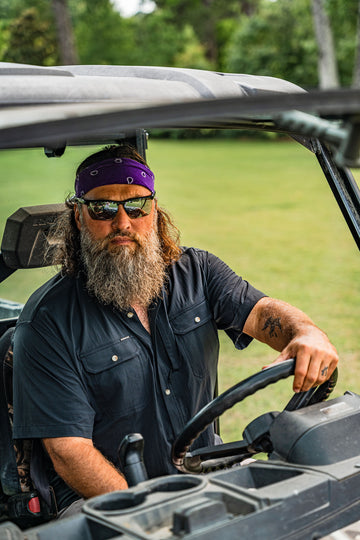 Bearded man wearing sunglasses and a purple bandana driving an off-road vehicle outdoors