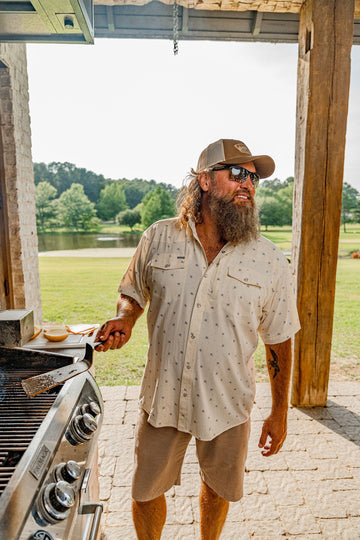 Man with beard grilling outdoors wearing a light patterned shirt and cap