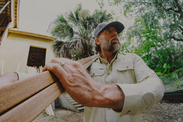 Man in outdoor shirt holding wooden planks near tropical trees and a building