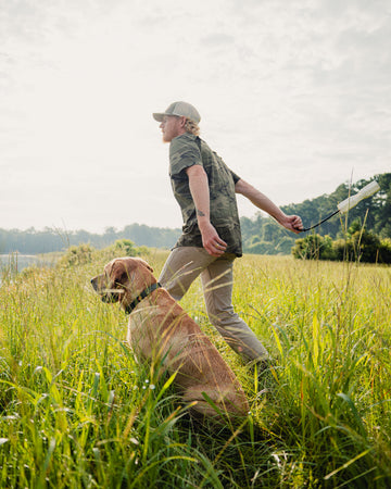 Young man in camo shirt walking dog through tall grass on a sunny day outdoors
