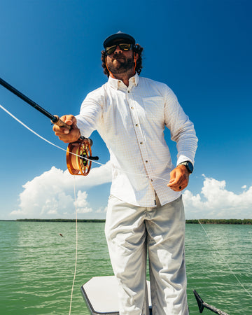 Man wearing sunglasses and a white long-sleeve shirt fly fishing on a boat under clear blue skies