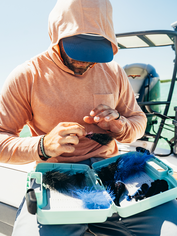 Man wearing a hooded shirt and cap tying fishing flies on a boat with a tackle box of colorful feathers