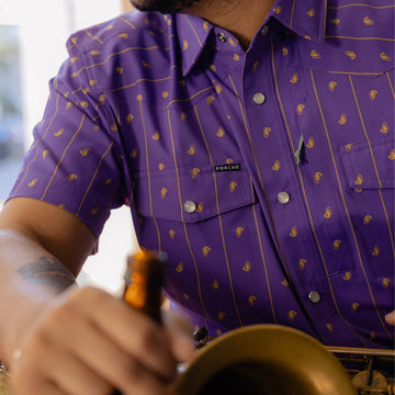 Man wearing The Hoodoo purple patterned outdoor shirt holding a saxophone and bottle