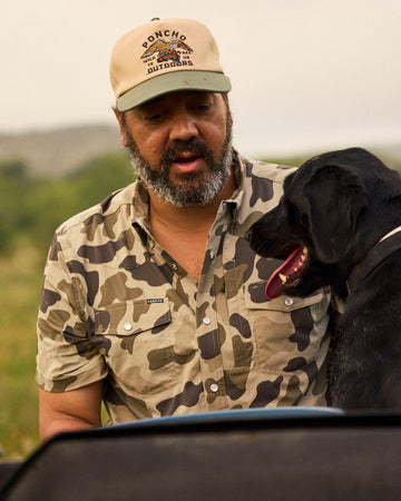 Man wearing a camo shirt and beige cap with a black dog outdoors