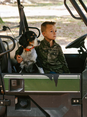 Boy in camouflage shirt sitting in vehicle with black and white dog wearing orange collar