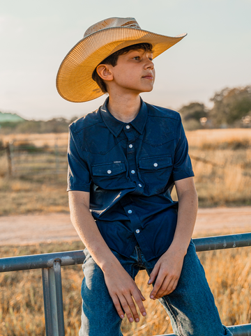 Boy wearing a cowboy hat and navy shirt sitting outdoors by a metal fence at sunset
