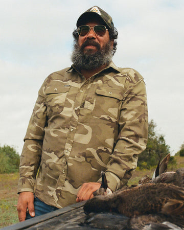 Man wearing camouflage shirt and hat standing outdoors next to hunting gear