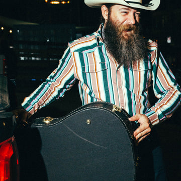 Man wearing The Frontman striped outdoor shirt and cowboy hat holding guitar case at night