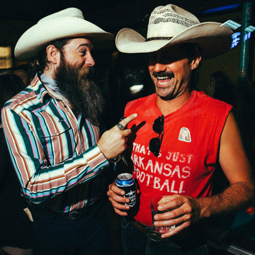 Two men wearing cowboy hats, one in a colorful striped shirt and the other in a sleeveless red shirt labeled The Frontman, laughing and holding drinks indoors