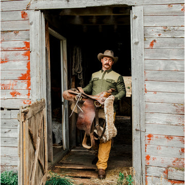 Man wearing The Roosevelt outdoor shirt holding a leather saddle in rustic barn doorway
