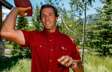 Man in red button-up shirt preparing to throw football outdoors in grassy area