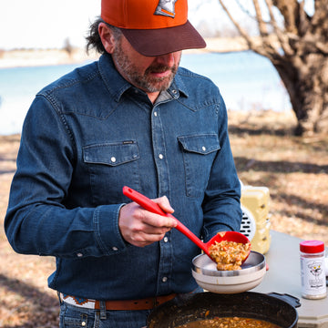 Man wearing The Sanderson denim outdoor shirt serving stew outdoors by a lake