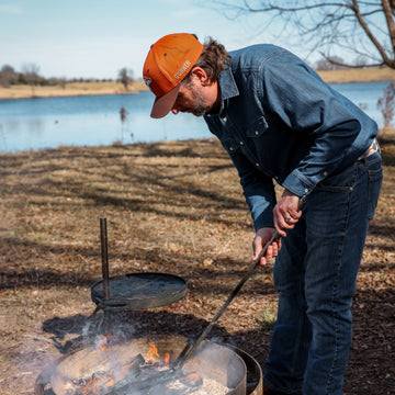 Man wearing The Sanderson denim shirt outdoors tending a campfire by a lakeside