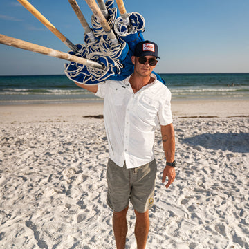 Man on beach wearing The Surf No Turf shirt carrying folded beach umbrellas on shoulder