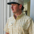Man wearing a light yellow plaid shirt called The Sweetwater looking outside near white columns