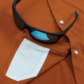 Black sunglasses resting on rust-colored shirt with white pocket, featuring The 1969 design
