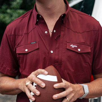 Man wearing a maroon "The Aggie" shirt holding a football outdoors