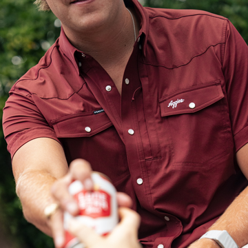 Man wearing a maroon shirt named The Aggie handing a can outdoors