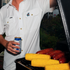 Person wearing The Albert white shirt grilling corn and sausages while holding a drink outdoors