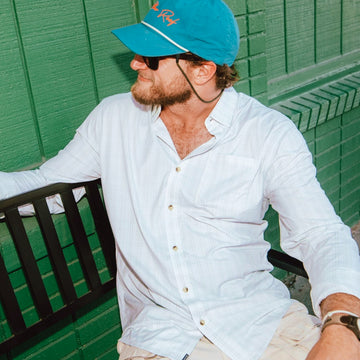 Man wearing The Destin white outdoor shirt sitting on bench by green wall