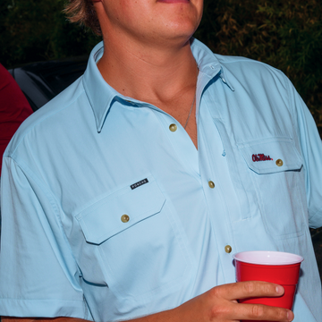Close-up of a light blue shirt from The Grove with buttoned pockets and a man holding a red cup outdoors