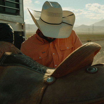 Man wearing a white hat and orange shirt outdoors with saddle in front titled The Joshua Tree