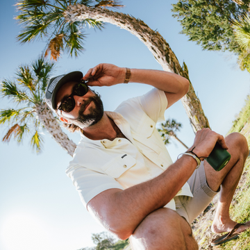 Man wearing "The Key West" shirt outdoors sitting on grass under palm trees in sunlight