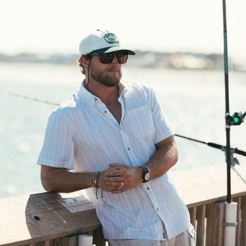 Man wearing The Seagrove Beach shirt leaning on wooden railing by water with fishing rods nearby