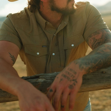 Man wearing The Sonora outdoors leaning on wooden fence with tattooed arms