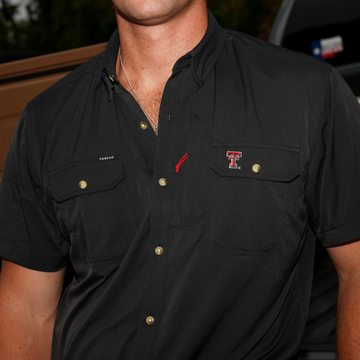 Close-up of a man wearing a black short-sleeve button-up shirt featuring The Tortilla logo on the chest pocket