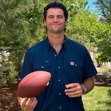 Man wearing The War Eagle shirt holding a football outdoors surrounded by greenery