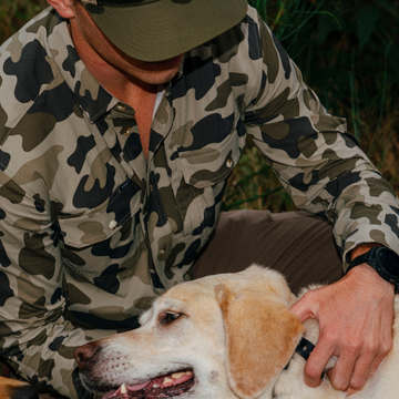 Man wearing The Zenaida camo shirt outdoors petting a yellow Labrador retriever dog