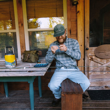 Man in The Cashiers shirt sitting on wooden bench tying fishing line outdoors