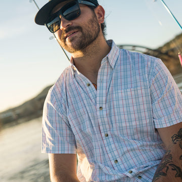 Man wearing The Mullet short-sleeve plaid outdoor shirt by water with sunglasses and hat