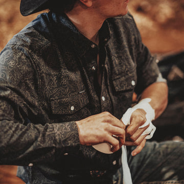 Man wearing The Pat Garrett patterned outdoor shirt and black hat wrapping his hands with white tape