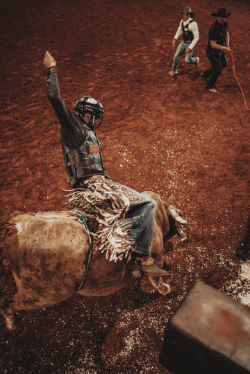 Rodeo rider in protective gear riding a bull on dirt arena with two cowboys in background