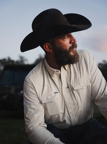 Bearded man wearing a black cowboy hat and beige outdoor shirt at dusk