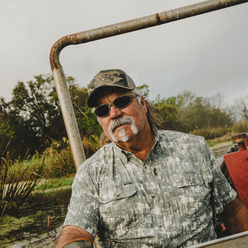 Man wearing sunglasses and a Camo Wild and Free Trucker Hat outdoors near swampy vegetation