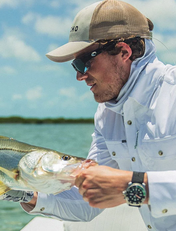 Man wearing Outdoors cap and white fishing shirt holding a fish near water