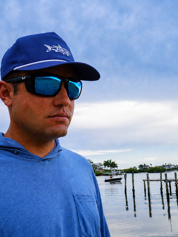 Man wearing blue shirt, sunglasses, and cap by waterfront with dock and boats under cloudy sky